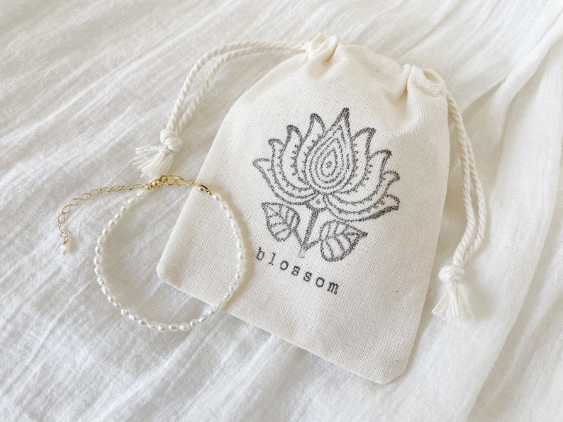 May include: A delicate pearl bracelet with a gold clasp, displayed on a white fabric pouch with a black floral design and the word "blossom" printed on it.