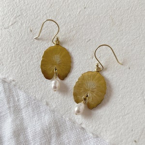 A pair of gold earrings with a pearl dangle. Each earring features a round, leaf-shaped design with a detailed etched pattern.