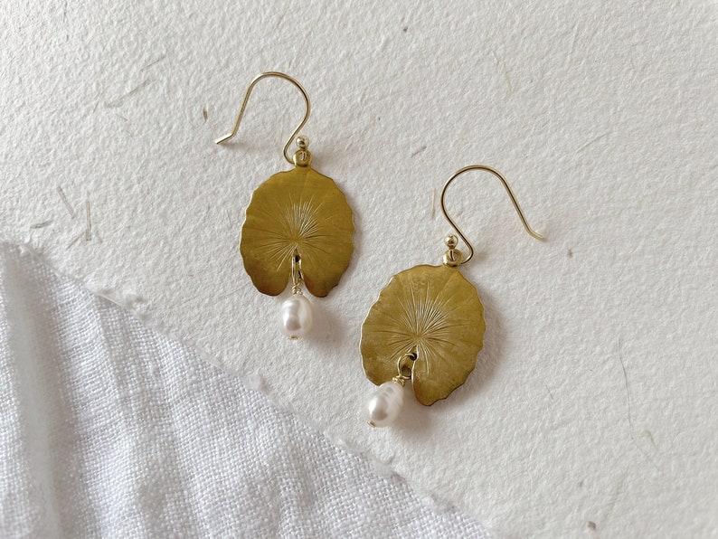 A pair of gold earrings with a pearl dangle. Each earring features a round, leaf-shaped design with a detailed etched pattern.