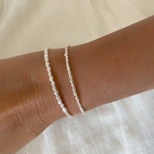 May include: Two delicate pearl bracelets on a wrist. The bracelets are made of small, white pearls strung together on a silver chain.