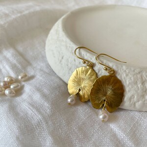 A pair of gold earrings with a leaf design and a pearl dangle. The earrings are on a white surface.