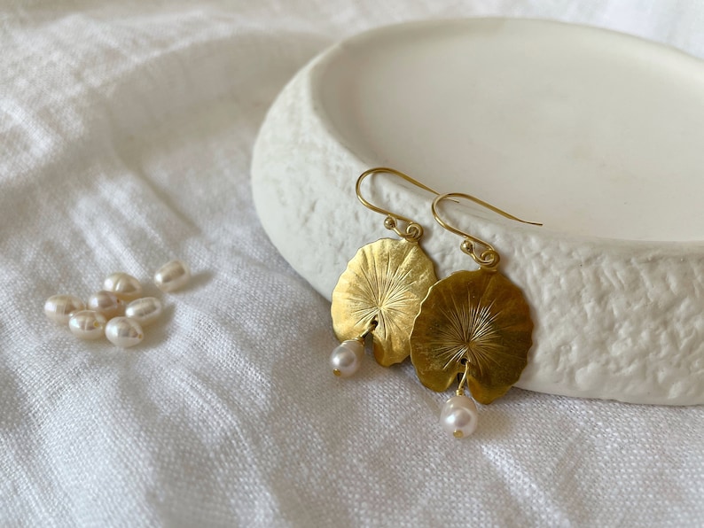 A pair of gold earrings with a leaf design and a pearl dangle. The earrings are on a white surface.