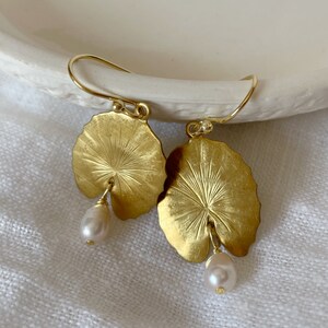 A pair of gold earrings with a leaf design and a pearl dangle. The leaves are textured and have a raised center. The small pearls are white and slightly round.