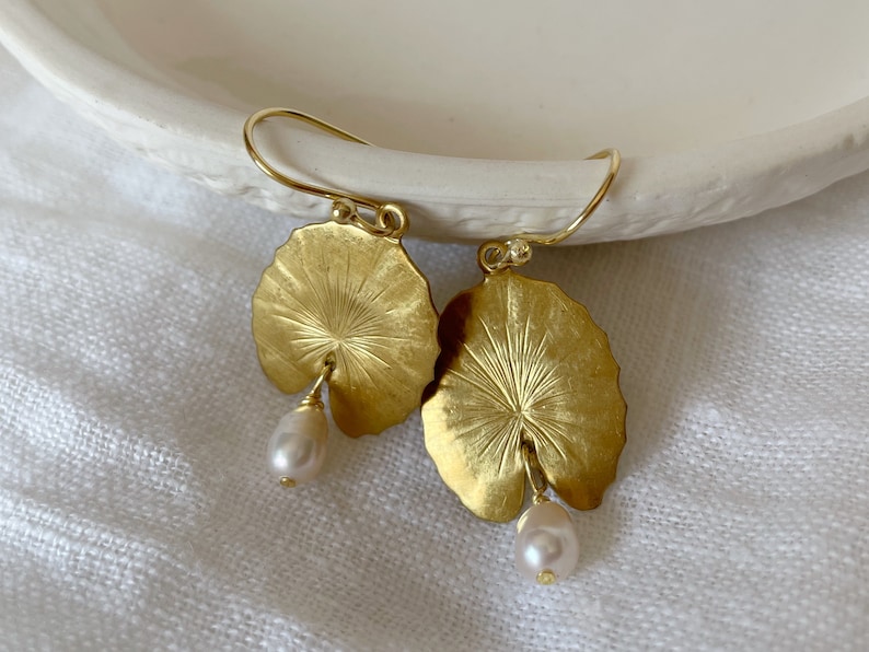 A pair of gold earrings with a leaf design and a pearl dangle. The leaves are textured and have a raised center. The small pearls are white and slightly round.