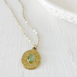 A gold necklace with a round pendant featuring a green gemstone. The pendant has a textured surface.