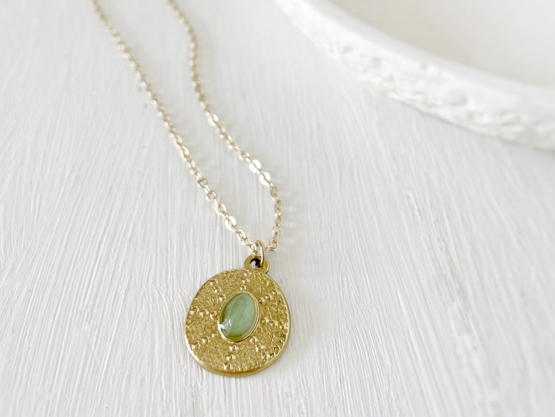 A gold necklace with a round pendant featuring a green gemstone. The pendant has a textured surface.