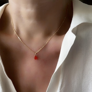 Può includere: Una delicata collana d'oro con un piccolo pendente a forma di goccia di pietra preziosa rossa. La collana è indossata su una camicia bianca, creando un look semplice ma elegante. La catena è sottile e il pendente è di un rosso vibrante.