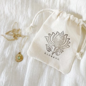 A gold necklace with a round pendant featuring a green stone. The necklace is in a small white drawstring bag with a black floral design and the word "blossom" printed on it.