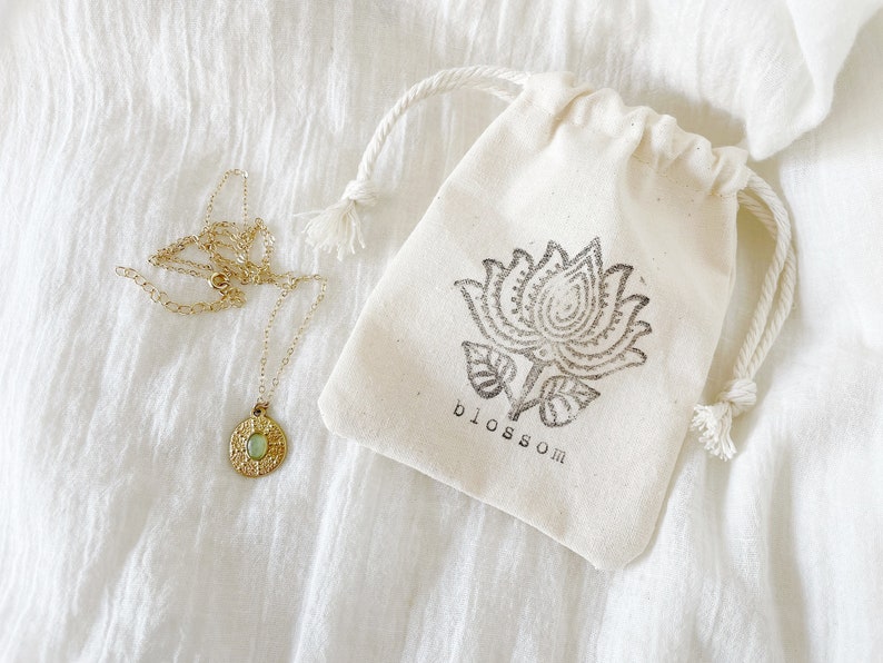 A gold necklace with a round pendant featuring a green stone. The necklace is in a small white drawstring bag with a black floral design and the word "blossom" printed on it.