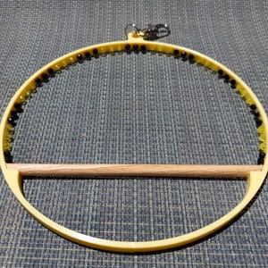 May include: A yellow bird toy with a wooden perch. The circular frame is adorned with black and yellow beads. A silver clasp is attached to the top for hanging. The toy is designed for small birds.