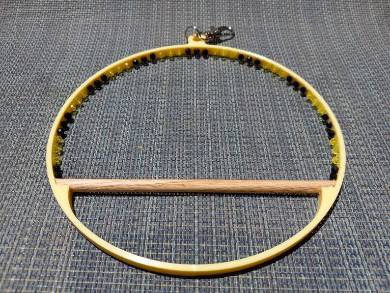 May include: A yellow bird toy with a wooden perch. The circular frame is adorned with black and yellow beads. A silver clasp is attached to the top for hanging. The toy is designed for small birds.