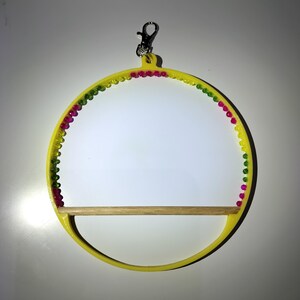 May include: A yellow bird perch with a wooden dowel and a decorative beaded trim in pink, green, and yellow. The perch has a silver clip for hanging. The design is circular.