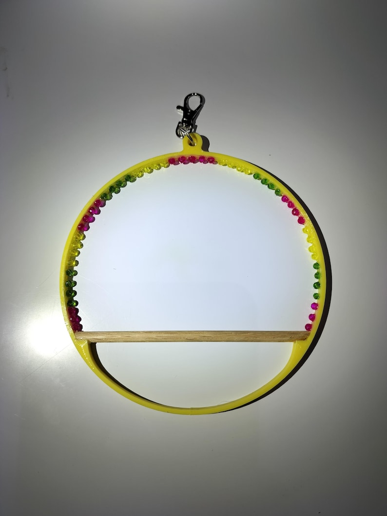 May include: A yellow bird perch with a wooden dowel and a decorative beaded trim in pink, green, and yellow. The perch has a silver clip for hanging. The design is circular.