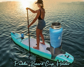 Może przedstawiać: Uchwyt na kubek do paddleboardu z dużą niebieską butelką na wodę. Uchwyt jest przymocowany do turkusowej deski do wiosłowania. Tekst "Paddle Board Cup Holder" jest napisany kursywą na dole obrazu.