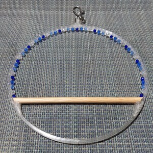 May include: Clear acrylic bird perch with a wooden dowel and a ring for hanging. The perch is decorated with blue and clear beads. A metal clasp is attached to the top for easy hanging. This bird perch is a decorative and functional accessory for pet birds.