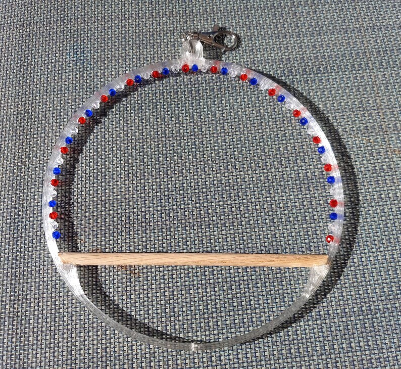 May include: Clear acrylic bird toy with red and blue beads around the perimeter. A natural wood perch is attached at the bottom. A silver-colored clasp is at the top for hanging.