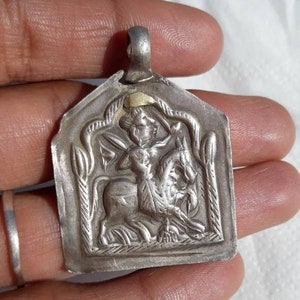May include: A silver pendant with a detailed depiction of a figure riding a horse. The pendant has a gold accent on the top and is in the shape of a shield.
