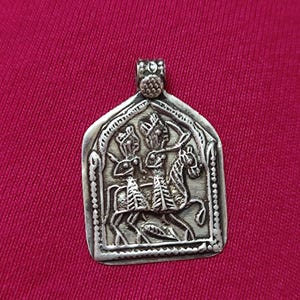 May include: Silver pendant with an engraved design of two figures, one on horseback, within an arched frame. The pendant has a detailed, antique appearance and is displayed against a red background.