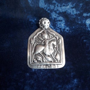 May include: A silver pendant with a detailed design of a person riding a horse. The pendant is surrounded by a decorative border and has a floral motif. The pendant is on a blue fabric background.