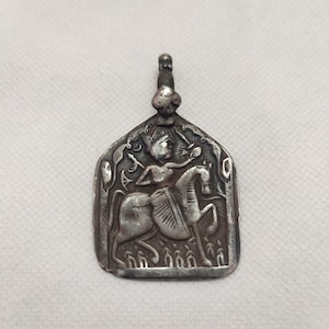 May include: A silver pendant featuring a detailed engraving of a figure riding a horse, holding a sword. The pendant has an ornate design with a loop at the top for a chain. The overall design is rectangular with rounded edges.