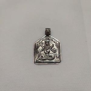 Könnte beinhalten: Silberanhänger mit geprägtem Design einer Gottheit. Der Anhänger hat eine rechteckige Form mit dekorativem Rand und einer Schlaufe für eine Kette. Die Gottheit wird mit mehreren Armen und einer schlangenartigen Form dargestellt.