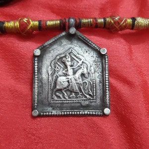 May include: A silver pendant with a detailed image of a person riding a horse. The pendant is attached to a black and red cord with gold accents.