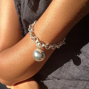 Peut inclure: Un bracelet en argent avec une grosse breloque ronde. Le bracelet est composé de maillons épais et imbriqués. Le charme est une sphère polie, reflétant la lumière. Le fond est un tissu sombre.