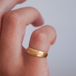 Puede incluir: Un anillo dorado con la palabra "promise" grabada en el exterior. El anillo se lleva en un dedo. El fondo está suavemente enfocado.