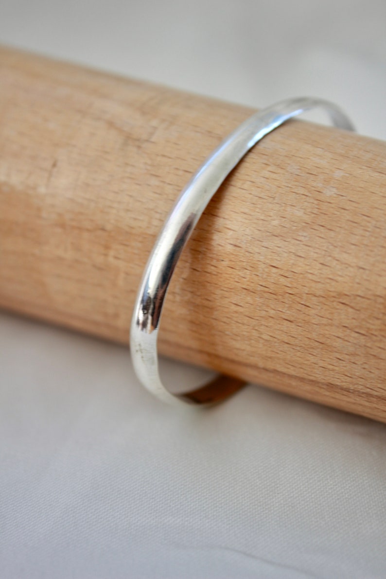 May include: A simple silver bangle bracelet, shown on a wooden surface.