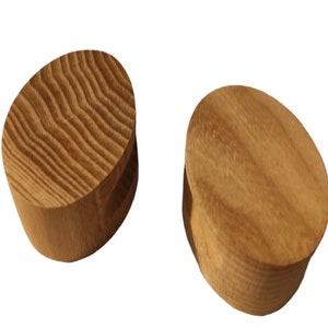 Puede incluir: Dos pomos de madera de forma ovalada con un acabado liso de color marrón claro. Los pomos tienen una parte superior ligeramente redondeada y una parte inferior plana.