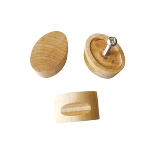 Puede incluir: Tres pomos de armario de madera sin terminar. Uno es de forma ovalada, otro es un óvalo plano con un tornillo, y el tercero es un pequeño cilindro con un óvalo empotrado. La madera tiene un color claro natural.