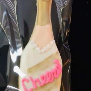 K&ouml;nnte beinhalten: Keks in Champagnerflaschenform mit goldener und wei&szlig;er Glasur. Der Keks hat eine rosa Aufschrift "Cheers" und ein Perlenhalsband-Detail. Der Keks befindet sich in einer transparenten Plastikt&uuml;te.