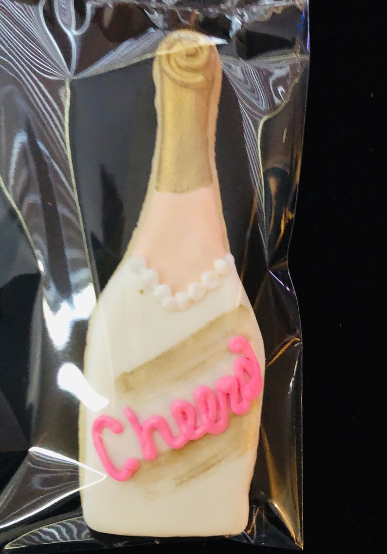 K&ouml;nnte beinhalten: Keks in Champagnerflaschenform mit goldener und wei&szlig;er Glasur. Der Keks hat eine rosa Aufschrift "Cheers" und ein Perlenhalsband-Detail. Der Keks befindet sich in einer transparenten Plastikt&uuml;te.