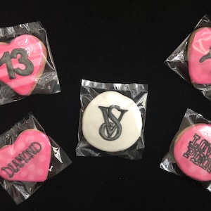 Könnte beinhalten: Auswahl an dekorierten Keksen in durchsichtiger Kunststoffverpackung. Herzförmige Kekse mit rosa Zuckerguss und schwarzem Text oder Designs, darunter die Zahl 13, das Wort "DIAMOND" und ein Logo. Ein runder Keks zeigt ein Logo.