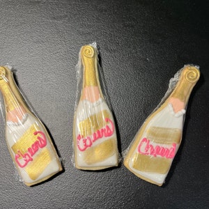 K&ouml;nnte beinhalten: Drei Sektflaschen-f&ouml;rmige Kekse, einzeln in Klarsichtfolie verpackt. Die Kekse sind mit goldener und wei&szlig;er Glasur verziert, mit dem Wort "Cheers" in Pink geschrieben. Die Kekse liegen auf einer dunklen Oberfl&auml;che.