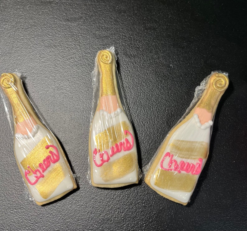 K&ouml;nnte beinhalten: Drei Sektflaschen-f&ouml;rmige Kekse, einzeln in Klarsichtfolie verpackt. Die Kekse sind mit goldener und wei&szlig;er Glasur verziert, mit dem Wort "Cheers" in Pink geschrieben. Die Kekse liegen auf einer dunklen Oberfl&auml;che.