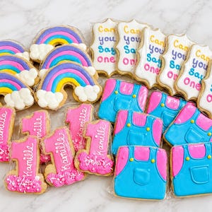 Könnte beinhalten: Eine Sammlung verzierter Kekse mit Regenbogenformen mit weißen Wolken, rechteckigen Keksen mit dem Text Can you say one, Keksen in Form der Zahl Eins mit dem Namen Kamille sowie blau-rosa Keksen in Form von Latzhosen.