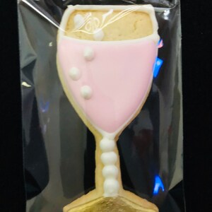 K&ouml;nnte beinhalten: Ein Keks in Champagnerglasform, verziert mit rosa Zuckerguss und wei&szlig;en Akzenten, ist in einer transparenten Plastikt&uuml;te versiegelt. Der Keks hat eine goldene Basis und einen Stiel mit wei&szlig;en perlenartigen Details. Das Glas ist mit einem hellen Keks gef&uuml;llt.