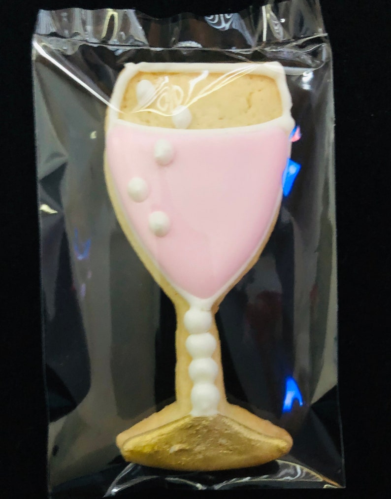 K&ouml;nnte beinhalten: Ein Keks in Champagnerglasform, verziert mit rosa Zuckerguss und wei&szlig;en Akzenten, ist in einer transparenten Plastikt&uuml;te versiegelt. Der Keks hat eine goldene Basis und einen Stiel mit wei&szlig;en perlenartigen Details. Das Glas ist mit einem hellen Keks gef&uuml;llt.
