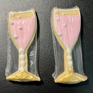 K&ouml;nnte beinhalten: Zwei Sektgl&auml;ser-f&ouml;rmige Kekse, einzeln in Klarsichtfolie verpackt. Die Kekse haben eine hellfarbene Basis mit einer rosa F&uuml;llung, wei&szlig;en Akzenten und goldfarbenen Basen. Die Kekse sind mit wei&szlig;en Blasendetails verziert.