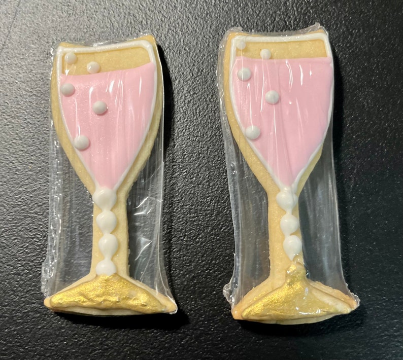 K&ouml;nnte beinhalten: Zwei Sektgl&auml;ser-f&ouml;rmige Kekse, einzeln in Klarsichtfolie verpackt. Die Kekse haben eine hellfarbene Basis mit einer rosa F&uuml;llung, wei&szlig;en Akzenten und goldfarbenen Basen. Die Kekse sind mit wei&szlig;en Blasendetails verziert.