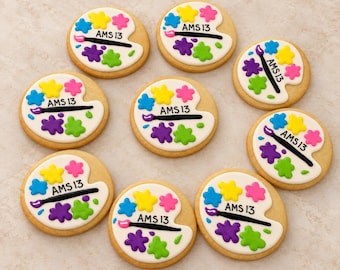 May include: Eight round sugar cookies decorated with white icing shaped like an artist's palette. Each cookie features colorful paint splatters in yellow, pink, blue, purple, and green, a black paintbrush, and the text AMS 13 in black icing.