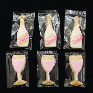 K&ouml;nnte beinhalten: Sechs einzeln verpackte Zuckerkekse. Drei sind wie Champagnerflaschen geformt, mit dem Wort "Cheers" in rosa Zuckerguss. Die anderen drei sind wie Champagnergl&auml;ser geformt, mit rosa F&uuml;llung und wei&szlig;en Punkten.