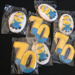 Könnte beinhalten: Kekse mit Minion-Charakteren und der Zahl 70 aus gelbem und blauem Zuckerguss verziert, einzeln in durchsichtigen Plastiktüten verpackt. Die Kekse liegen auf einer dunklen Oberfläche.