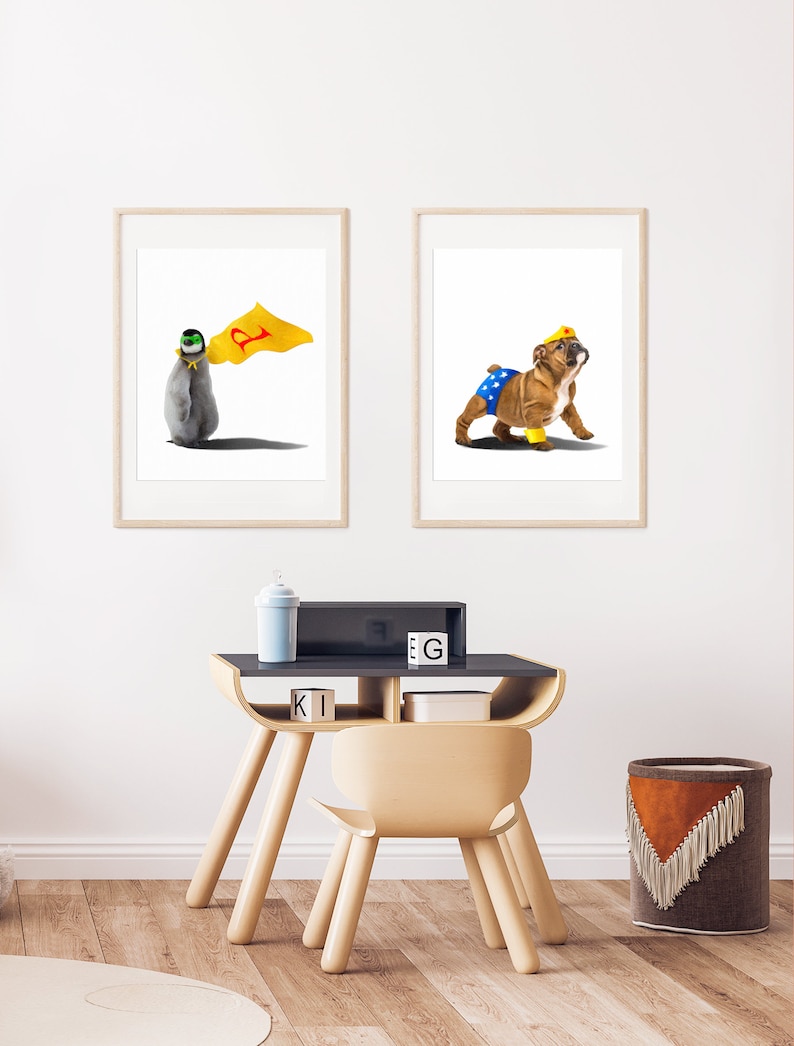 May include: Two framed illustrations of animals dressed as superheroes. The first image shows a penguin wearing a yellow cape with a red letter 'P' on it. The second image shows a bulldog wearing a blue and yellow Wonder Woman costume. The illustrations are displayed on a white wall above a wooden desk with a chair.