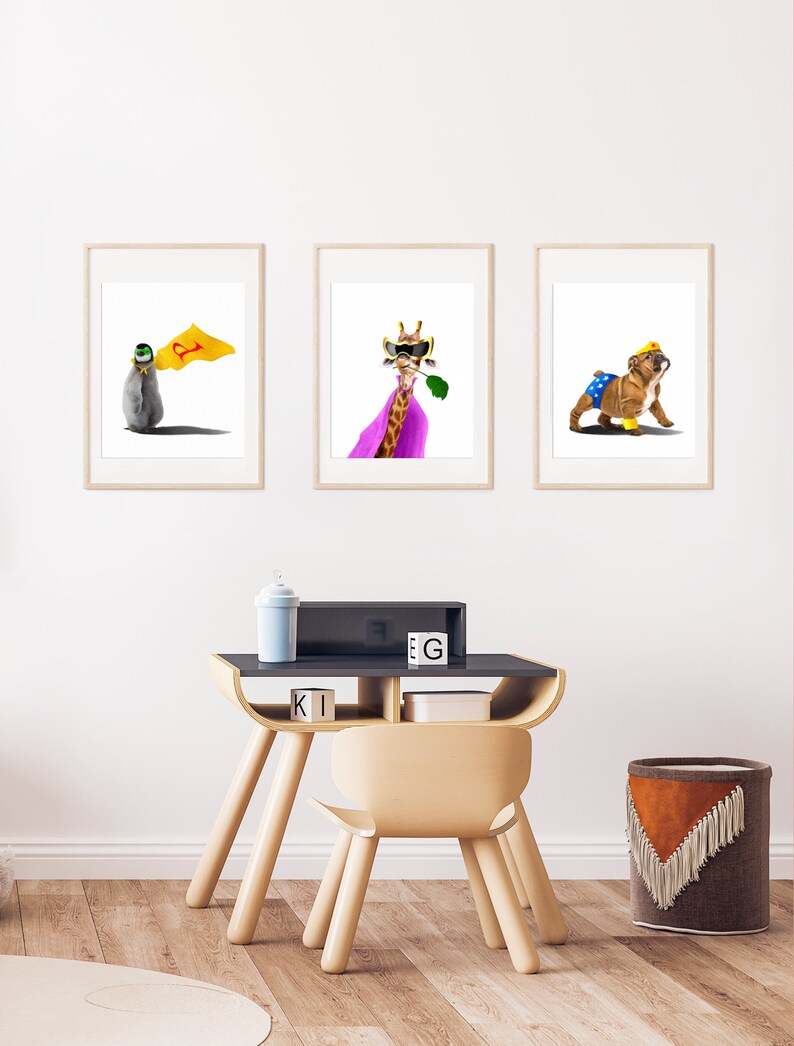 May include: Three framed animal illustrations in a children's room. The illustrations feature a penguin wearing a cape, a giraffe wearing a cape and sunglasses, and a bulldog wearing a cape and a crown. The illustrations are hanging on a white wall above a wooden desk and chair.