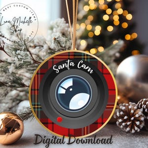 Op de afbeelding: Een ronde kerstbal met een rood-zwart ruitpatroon. De bal heeft een camera lens met de tekst "Santa Cam" in een witte cirkel.
