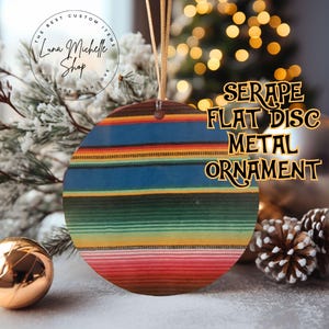 May include: A round metal ornament with a serape design in red, blue, green, and yellow stripes. The ornament has the text "SERAPE FLAT DISC METAL ORNAMENT". It is hanging by a gold string. The Luna Michelle Shop logo is in the background.
