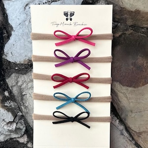 May include: A set of five baby headbands with small suede bows in pink, purple, red, teal, and black. The headbands are on a cream-colored card with the text "Tiny Miracle Bowties" and a footprint logo.