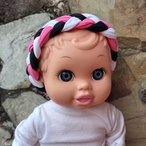 May include: A doll with blue eyes and a pink, white, and black braided headband. The doll is wearing a white shirt.